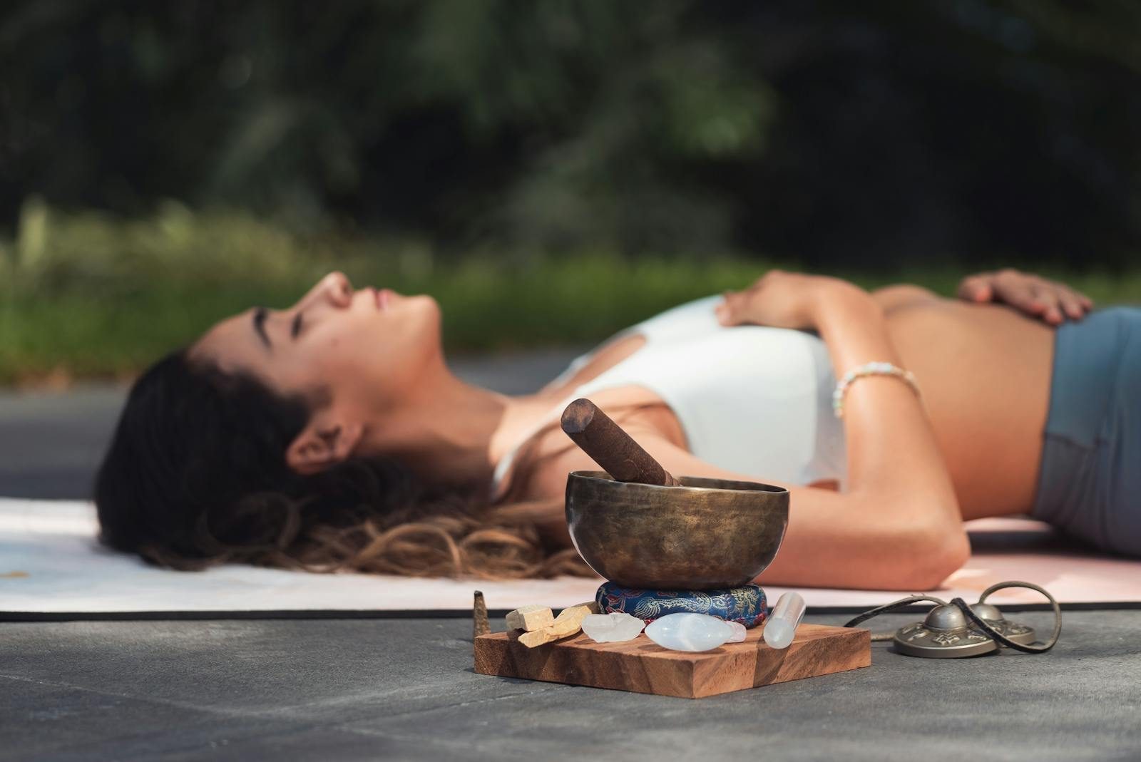 A woman practices relaxation with a singing bowl and crystals in an outdoor setting.